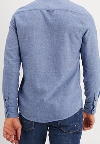Camicia a maniche lunghe blu, tessuto testurizzato, polsini con bottoni, orlo arrotondato, dettaglio della spalla posteriore, abbinata a jeans scuri.