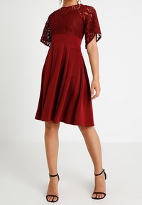 Burgundy klänning med spetsöverdrag, korta ärmar, figurnära liv och utsvängd kjol. Har veck och en slät textur. Svarta klackar med ankelsnörning.
