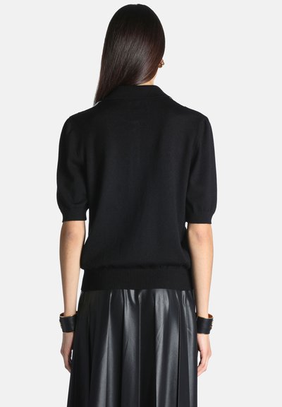Femme aux longs cheveux foncés portant un pull noir à manches courtes et une jupe plissée en cuir noire, debout de dos.