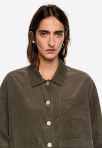Oliwkowa koszula kurtka z kołnierzem, zapinana na guziki z przodu, z kieszenią na piersi. Wykonana z fakturowanej tkaniny o casualowym kroju.