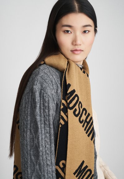 Jovem mulher com cabelo escuro e comprido usa um suéter cinza de malha em cordas e um lenço bege com texto preto, olhando calmamente para a câmara.