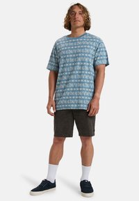 Blue patterned t-shirt featuring a leaf design, paired with dark shorts. Model wearing white socks and navy sneakers.