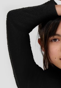 Zwarte hoge hals top van textuurstof. Lange mouwen met ruwe zomen. Model met donker haar gedeeltelijk zichtbaar, poseert met één arm omhoog.