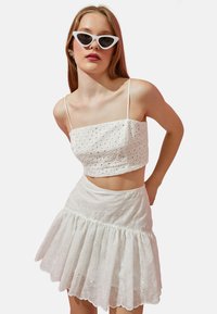 Top corto blanco de ojales con tirantes finos, combinado con una falda blanca acampanada y de capas. Presenta patrones florales y un dobladillo con forma de ondas.