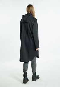 Cappotto nero con cappuccio, dalla vestibilità lunga e ampia, realizzato in tessuto testurizzato. Il retro presenta un orlo curvo, abbinato a pantaloni grigi e stivali neri spessi.