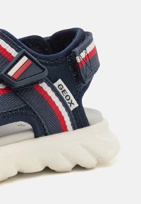 Navy blue sandal with a textured fabric upper, red and white striped accents, adjustable strap, and a chunky white rubber sole.