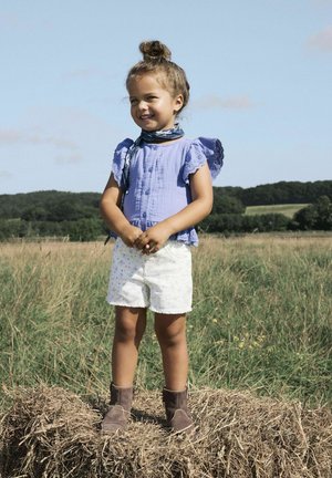 Top de manga corta lila claro con detalles de ojales, pantalones cortos blancos con estampado floral y botas marrones hasta el tobillo. De pie sobre heno en un campo.