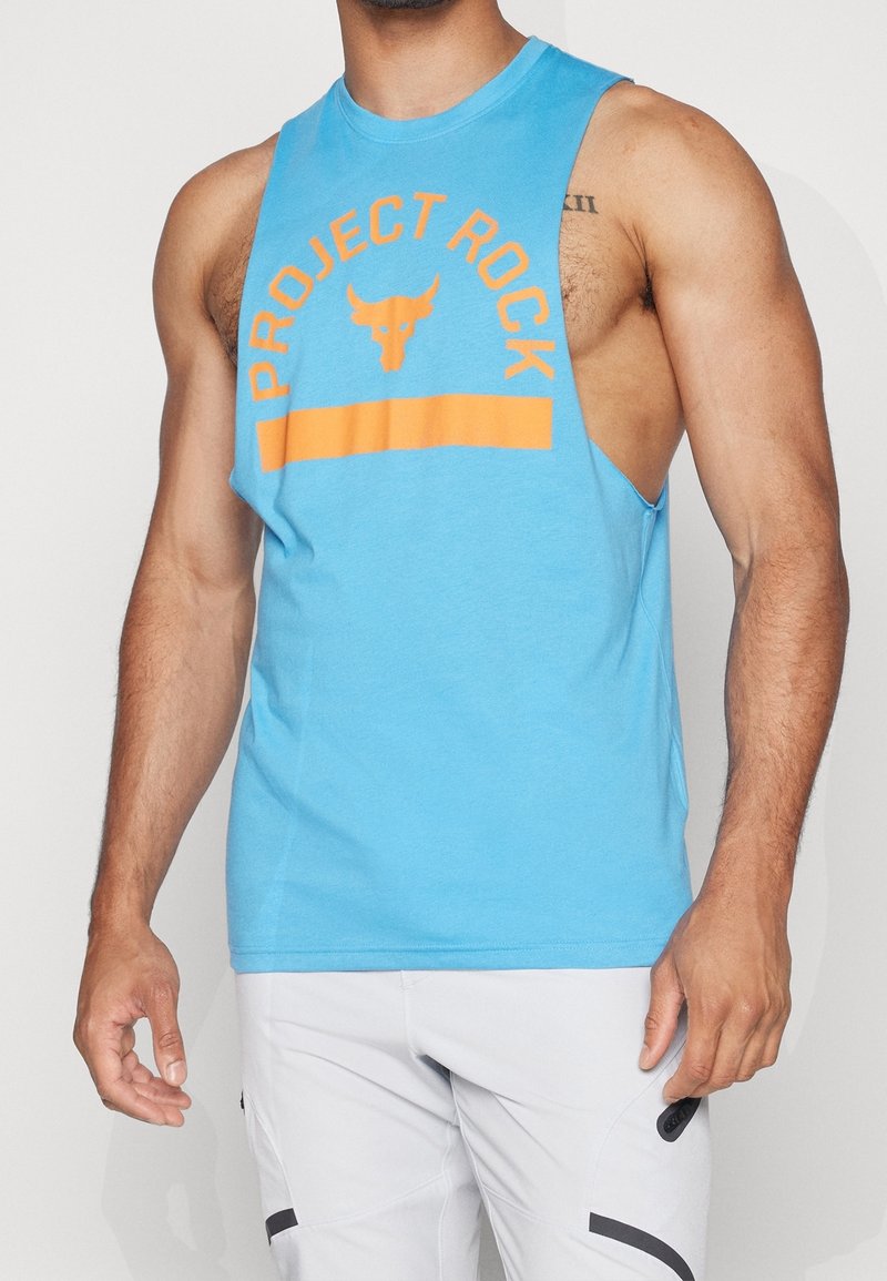 Homme portant un débardeur bleu vif avec texte orange "Project Rock" et logo de taureau, associé à un pantalon de sport gris clair.