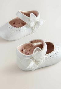 Baltās ādas Mary Jane kurpes ar viļņotu malu un zīda tauriņa akcentu uz siksniņas. Mīkste rozā iekšējā odere.