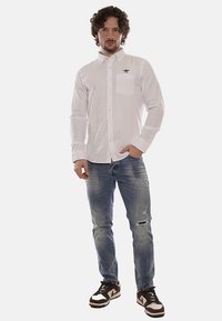Camicia bianca a maniche lunghe con bottoni, dotata di taschino e logo, abbinata a jeans blu strappati e sneakers marroni, in piedi contro uno sfondo neutro.
