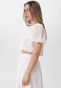 Biały krótki top wykonany z lekkiego materiału, z bufiastymi rękawami i wycięciami laserowymi, prezentujący teksturowany, oddychający design.