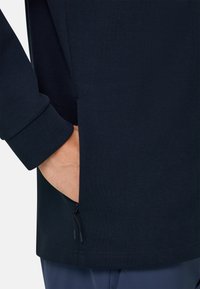 Hand in der Seitentasche einer marineblauen, mit Reißverschluss versehenen Jacke aus strukturiertem Stoff, kombiniert mit blauer Hose.