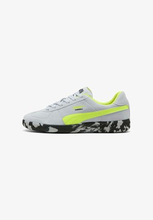 Zapatilla de balonmano Puma gris claro con franja interior de color amarillo neón, suela con patrón en blanco y negro, diseño de corte bajo con cordones.