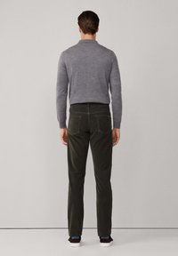 Man in een grijze trui met lange mouwen, donkere groene broek en donkere sneakers met witte zolen, staande met zijn gezicht naar een effen lichte achtergrond.