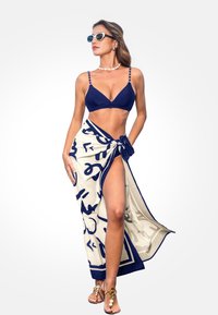 Marineblaues Bikini mit einem dreieckigen Oberteil, kombiniert mit einem cremefarbenen Wickeltuch mit marineblauen abstrakten Mustern. Das Modell trägt weiße Sonnenbrillen.