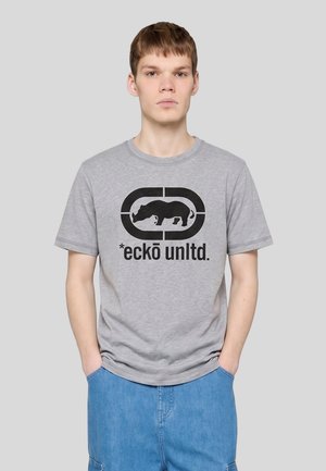 Jeune homme à la peau claire et aux cheveux bruns courts portant un t-shirt gris Ecko Unltd. avec un logo rhinocéros noir et un pantalon bleu, debout devant un fond uni.
