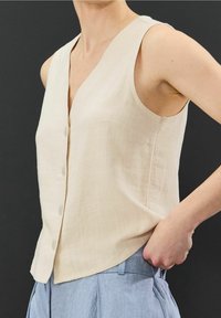 Blusa sin mangas beige hecha de tela texturizada, con escote en V y seis botones, combinada con pantalones azul claro.