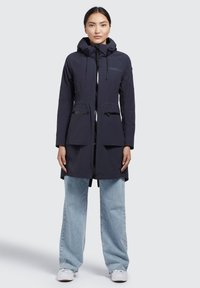 Cappotto blu navy con cappuccio, due tasche frontali, chiusura con zip e una forma leggermente sagomata; indossato sopra jeans a gamba larga blu chiaro.