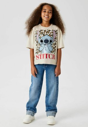 T-shirt beige avec un design graphique de Stitch sur fond léopard. Associé à un jean bleu ample et des baskets blanches.