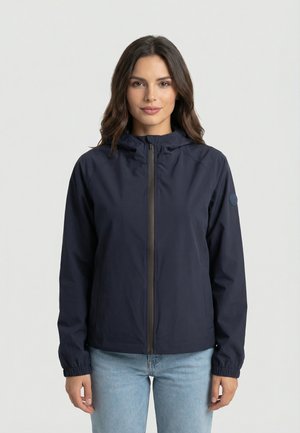Donna che indossa una giacca blu navy con zip e cappuccio e jeans azzurri, in piedi contro uno sfondo chiaro e uniforme, rivolta verso avanti.