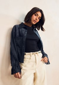 Chemise boutonnée en daim bleu marine avec des détails à franges aux manches, associée à un haut noir ajusté et un pantalon beige taille haute.