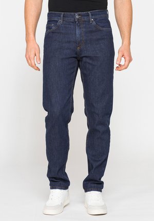 Uomo che indossa jeans blu scuro a gamba dritta e sneakers bianche, in piedi davanti a uno sfondo semplice.