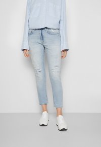 Vaqueros de mezclilla azul claro con un ajuste entallado, que presentan desgastes cerca de las rodillas. Combinados con una camisa abotonada azul pálido y zapatillas blancas.