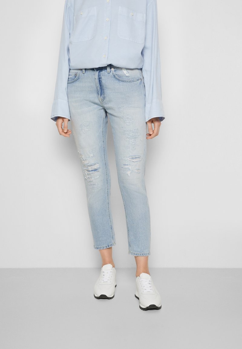 Vaqueros de mezclilla azul claro con un ajuste entallado, que presentan desgastes cerca de las rodillas. Combinados con una camisa abotonada azul pálido y zapatillas blancas.