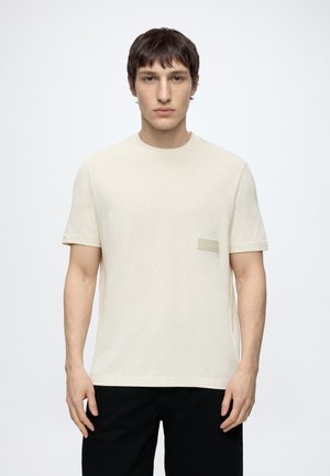 Jeune homme aux cheveux courts et foncés portant un t-shirt beige uni à manches courtes et un pantalon noir, debout devant un fond blanc.
