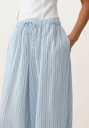 Žena nosi svetlomodré široké nohavice s vysokým pásom, tenkými vertikálnymi pruhmi a sťahovacou šnúrkou, ruka v vrecku, v kombinácii s bielym tielkom bez rukávov.