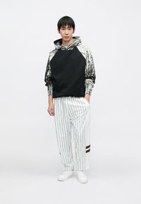 Czarna bluza z kapturem z wzorem i kangurzą kieszenią, zestawiona z białymi spodniami w cienkie prążki z bordowymi akcentami i mankietami, noszona z białymi butami.