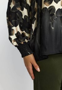 Hånd hviler ved siden, iført langermet bluse med svart blomstertrykk og rynkede mansjetter, kombinert med olivengrønne bukser.