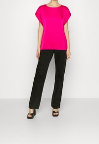 Una persona che indossa una blusa corta di un brillante colore rosa, pantaloni neri a gamba dritta e sandali con tacco aperti neri, in piedi contro uno sfondo bianco.