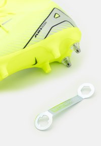 Cizma de fotbal galben strălucitor cu suprafață texturată și talpă cu cuie, asociată cu o cheie metalică argintie cu deschideri hexagonale.