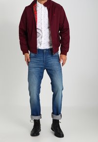 Cazadora burdeos sobre una camisa blanca de botonadura con un patrón, combinada con jeans azules y botas de cordones negras. La cazadora presenta un forro de cuadros.