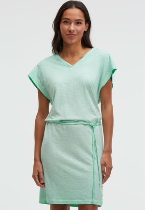 Frau trägt ein hellgrünes, kurzärmeliges Kleid mit Gürtel und V-Ausschnitt, steht vor einem einfarbigen hellen Hintergrund.