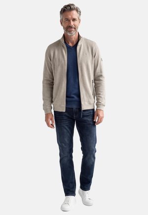 Mittelalter Mann mit grauen Haaren, der eine beige Jacke mit Reißverschluss, einen dunkelblauen Pullover, dunkle Jeans und weiße Turnschuhe trägt und vor weißem Hintergrund steht.