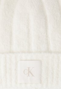 Bonnet en tricot blanc fabriqué à partir de tissu texturé doux, affichant un design côtelé et un petit écusson de couleur claire avec "cK" brodé dessus.