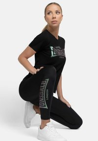 Black t-shirt with reflective "Lonsdale London" text, paired with black leggings featuring a logo accent. Worn with white sneakers.