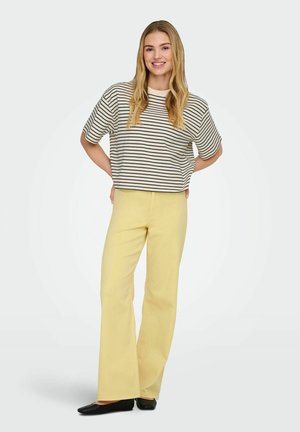 Usmievajúca sa žena s dlhými blond vlasmi, oblečená v čierno-bielom prúžkovanom tričku s krátkym rukávom, žltých nohaviciach a čiernych plochých topánkach.