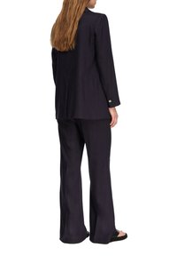 Donna con capelli lunghi che indossa un blazer nero e pantaloni larghi coordinati, in piedi a piedi nudi di spalle su uno sfondo bianco.