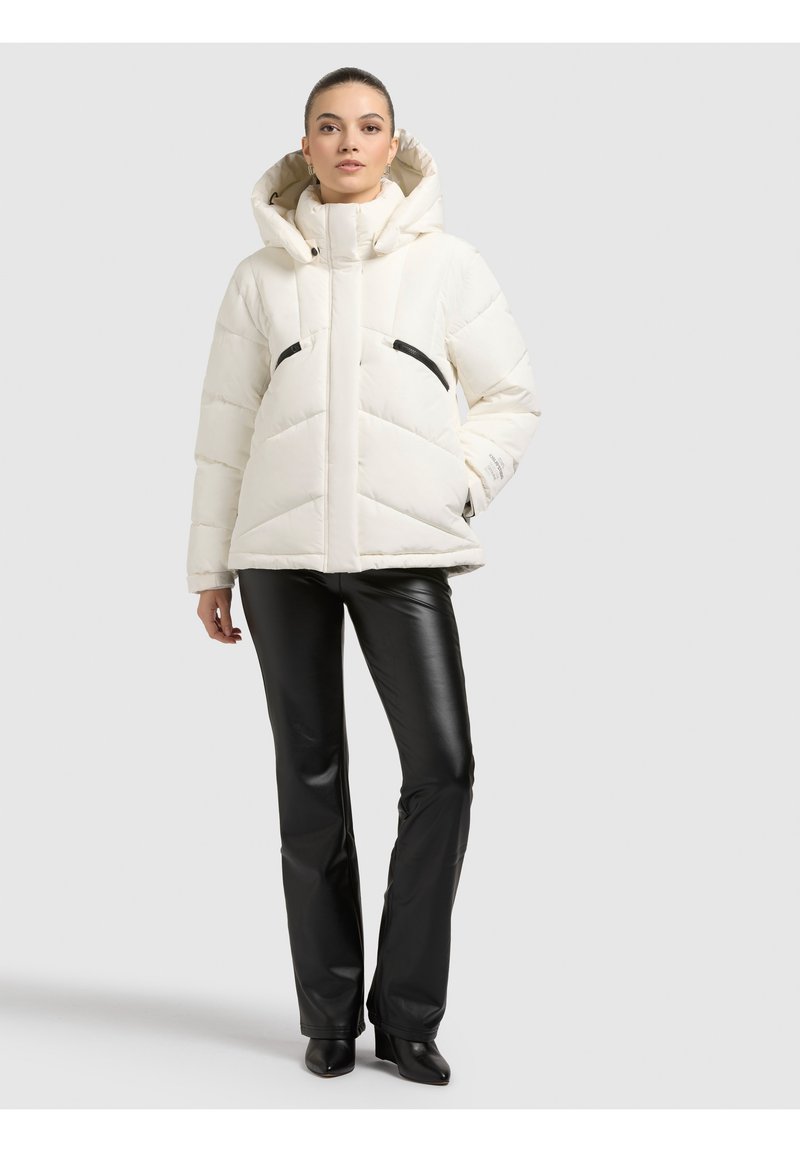 Biała puchowa kurtka z kapturem, z wzorem jodełki i czarnymi kieszeniami na zamek, zestawiona z czarnymi skórzanymi spodniami i botkami do kostki.