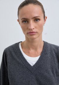 Jeune femme aux cheveux tirés en arrière, portant des boucles d'oreilles en argent et un pull gris à col en V sur un haut blanc, devant un fond clair uniformément.