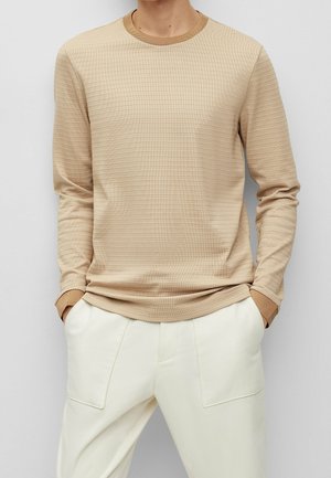 Mand iført beige ribstrikket langærmet sweater med kontrastfarvet krave og manchetter, kombineret med off-white bukser, hænderne i lommerne.
