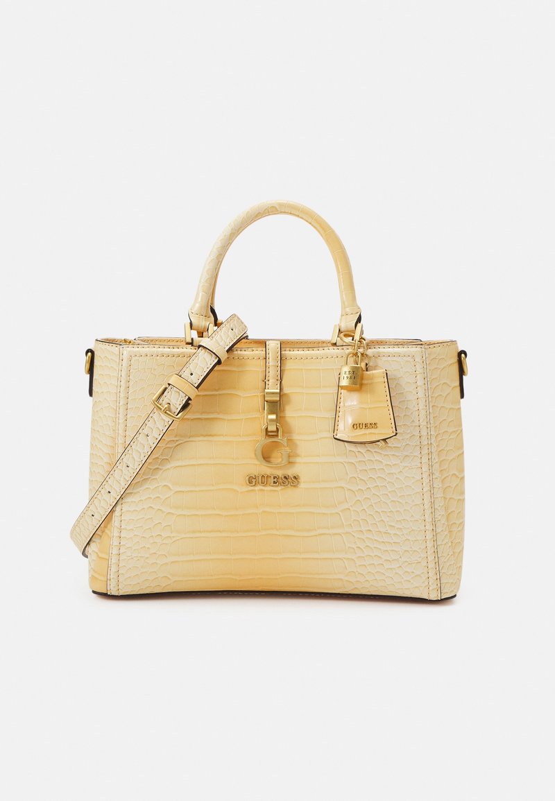 Sac à main en cuir beige embossé crocodile avec poignées supérieures, bandoulière ajustable, logo Guess doré et charm décoratif.