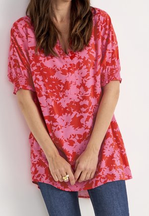 Kobieta ubrana w różowo-czerwoną, kwiecistą bluzkę z bufkami o długości do łokcia i niebieskie dżinsy, trzymająca ręce złożone blisko brzegu bluzki.