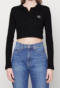 Svart långärmad kort polo med Calvin Klein-logotyp, ihop med högmidjade blå denimjeans på en stående person.