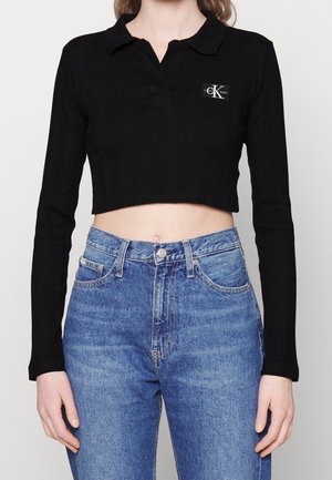 Schwarzes, langärmeliges, verkürztes Polo-Shirt mit Calvin Klein-Logo, kombiniert mit hoch taillierten blauen Jeans an einer stehenden Person.
