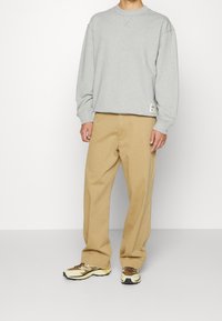 Felpa grigia con scollatura rotonda, maniche lunghe e etichetta sull'orlo; abbinata a pantaloni beige dalla vestibilità ampia e sneakers marroni e gialle.