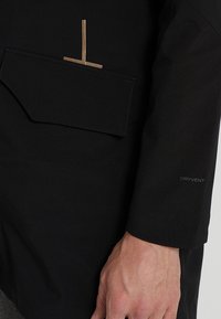 Giacca nera impermeabile con un tessuto testurizzato, un audace design delle tasche e un logo "DRYVENT" in evidenza sulla manica.